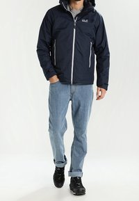 Mörkblå vattentät jacka med dragkedja framtill, grå detaljer, justerbar huva och två sidofickor, som bärs över ljusblå jeans och svarta skor.