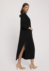 Długi czarny płaszcz o dopasowanym kroju, z bocznymi rozcięciami i przednimi kieszeniami. Posiada złote akcenty w postaci guzików i gładką fakturę. Nosisz go na beżowej odzieży.