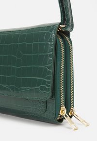 Borsa in pelle verde con effetto coccodrillo, dotata di chiusura a patta e due scomparti con zip. L'hardware dorato enfatizza il design.