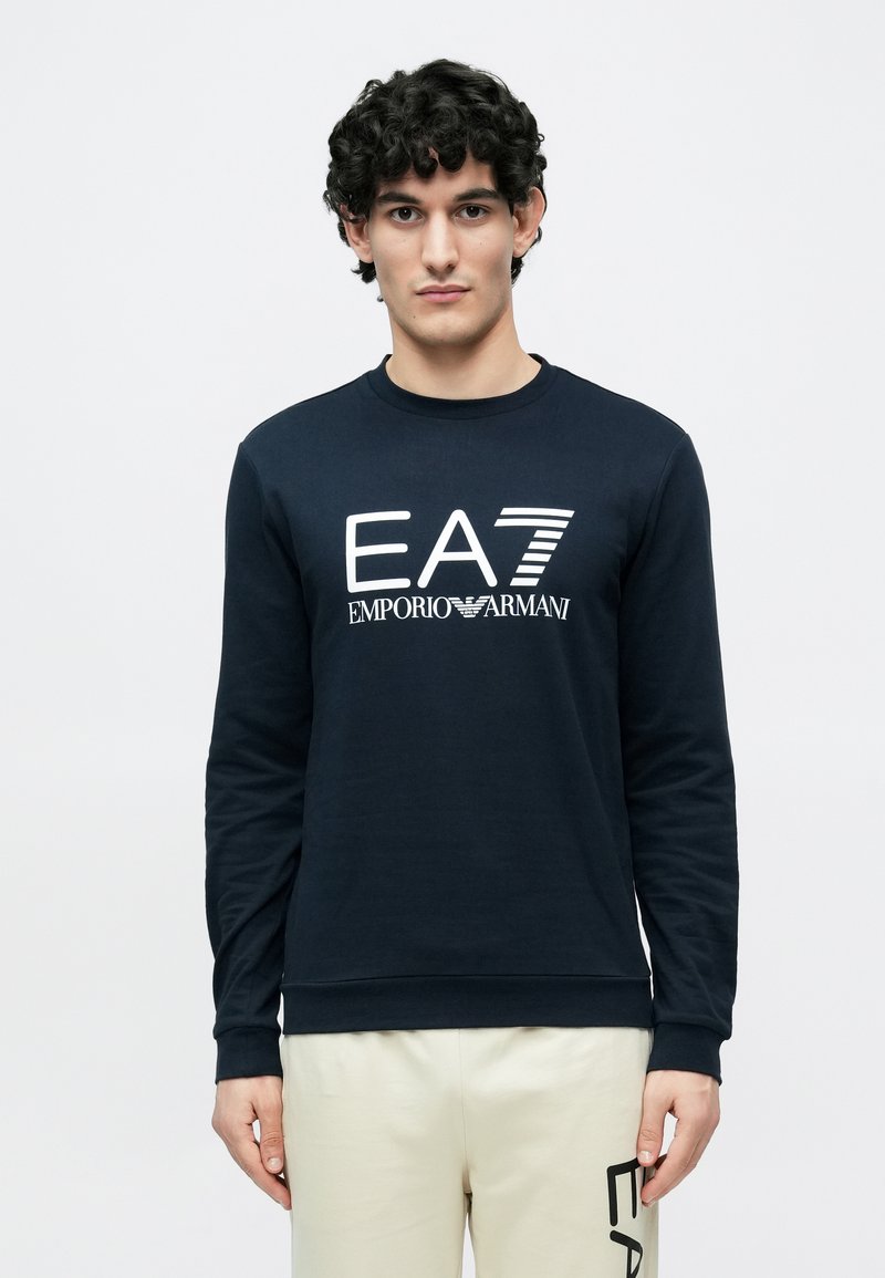 Temno modra jopa z dolgimi rokavi in rebrastimi manšetami, na sprednji strani z velikim belim logotipom "EA7 Emporio Armani". Narejena iz mehkega bombažnega materiala.