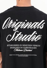 Black cotton t-shirt featuring white text in various fonts. Text includes "Originals Studio" and design details about the brand and location.