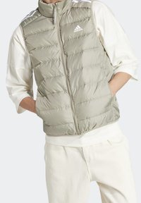 Pohodlná vesta v světlé olivově zelené barvě, vyrobená z lesklého materiálu se zipovým zapínáním a vysokým límcem. Obsahuje bílé logo Adidas a kontrastní pruhy.