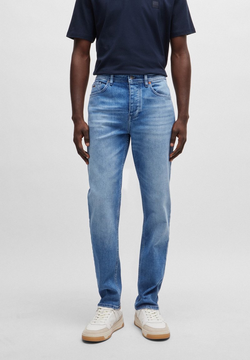 Man med urblekta blå jeans, marinblå t-shirt och vita sneakers som står mot en enkel ljus bakgrund.