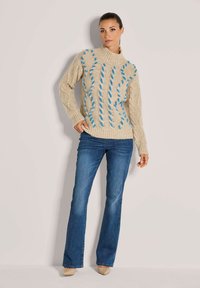 Frau in beigem grob gestricktem Pullover mit blauen Garnakzenten, blauen Bootcut-Jeans und beigen spitzen Schuhen, die vor einer weißen Wand steht.