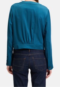 Teal cropped jacket with long sleeves, elastic waistband, and cuff detailing; paired with dark blue jeans featuring a classic pocket design.