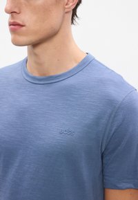 Svetlomodré tričko s krátkymi rukávmi vyrobené z mäkkého materiálu. Má okrúhly výstrih a jemne embosované logo "BOSS" na hrudi.