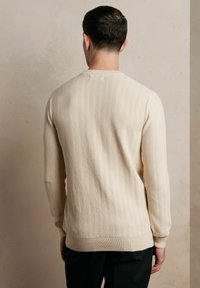 Dzianinowy sweter w kremowym kolorze z długim rękawem, wyróżniający się teksturowanym wzorem w rybie łuski oraz ściągaczami przy mankietach, dolnym brzegu i dekolcie, noszony na odwrót.