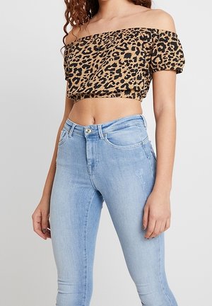 Brązowy i czarny crop top w panterkę z odkrytymi ramionami, w połączeniu z jasnoniebieskimi obcisłymi jeansami z zapięciem na guziki i przednimi kieszeniami.