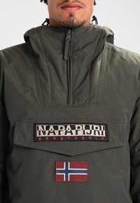 Olivengrøn anorakjakke med høj krave, frontlomme og broderede logoer, herunder et norsk flag-patch. Glat tekstur.