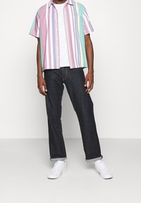 Uomo con una camicia a maniche corte a righe verticali multicolori, una maglietta bianca sotto, jeans scuri con risvolto e sneaker bianche, in piedi su uno sfondo semplice.