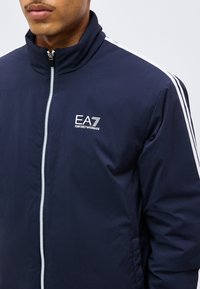 Άνδρας που φοράει navy μπλε Emporio Armani EA7 ζακέτα με φερμουάρ, με λευκές ρίγες στους ώμους και λευκό λογότυπο στο στήθος.