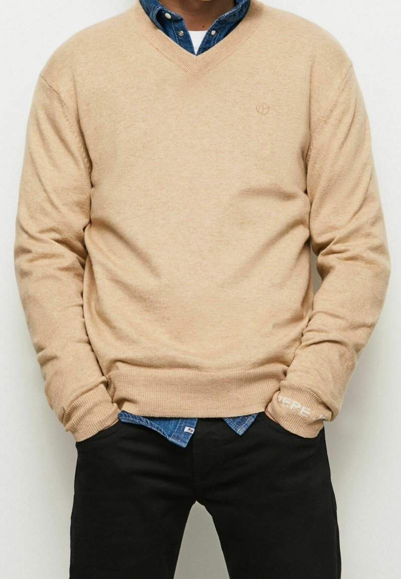 Homme portant un pull beige à col en V par-dessus une chemise bleue boutonnée, pantalon noir et mains dans les poches sur un fond uni.