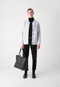 Biely sako s button-up zapínaním, čierny rolák, čierne nohavice a čierne topánky. Drží veľkú čiernu koženú tašku s embosovaným logom.