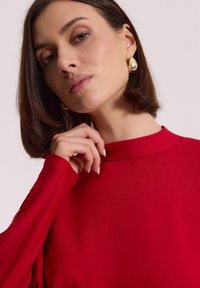 Kobieta z krótkimi brązowymi włosami, nosząca złote kolczyki w kształcie koła i czerwoną bluzkę z długim rękawem, delikatnie dotykająca swojej szyi na neutralnym tle.