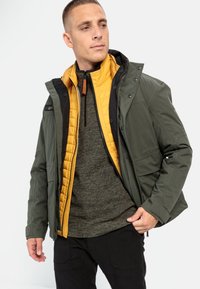 Lagdelte jakke med et olivengrønt yderlag, en quiltet sennepsgul foring og en tekstureret mørkegrå sweater under.