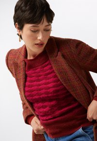 Strickpullover mit Struktur in tiefrot mit wellenförmigem Muster, darunter ein gemusterter Blazer in Rosa- und Bordeauxtönen, mit lässiger Passform.