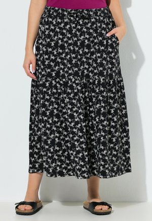 Kobieta ubrana w czarną spódnicę midi z białymi abstrakcyjnymi wzorami linii, do tego czarne klapki typu slide i fioletowa bluzka, stojąca przy białej ścianie.