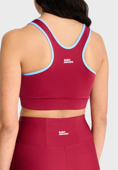 Femme portant un soutien-gorge de sport dos nageur bordeaux avec des bordures bleu clair et un legging taille haute assorti, vue de dos avec les logos des marques visibles.