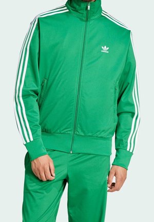 Mann trägt einen grünen Adidas-Trainingsanzug mit weißen Streifen an Ärmeln und Hosen, Reißverschlussjacke und Adidas-Logo auf der Brust und am Oberschenkel.