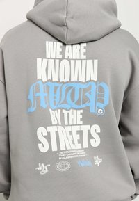 Sudadera con capucha gris con un gran texto gráfico en blanco y azul en la parte trasera que dice "WE ARE KNOWN MUTED BY THE STREETS" con logotipos adicionales debajo.