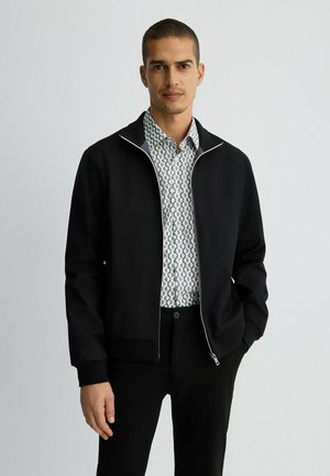 Black zip-up jacket with ribbed cuffs and collar, worn over a patterned white shirt. Features a sleek, lightweight design.