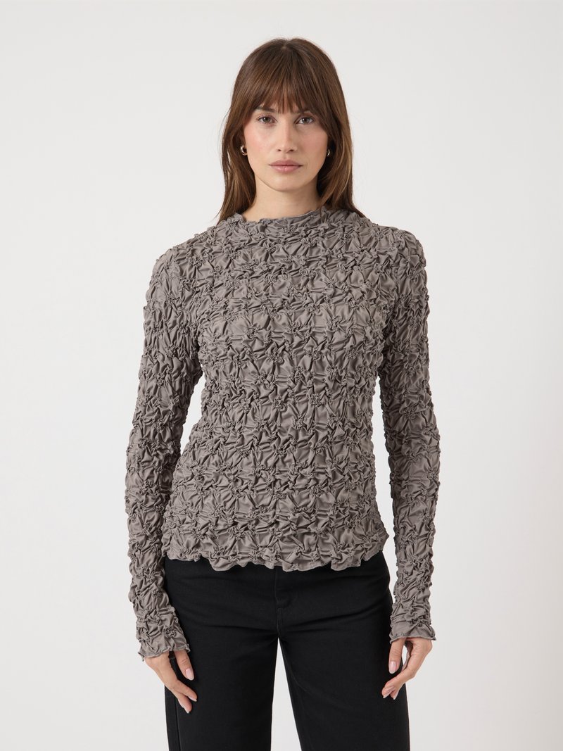 Femme aux cheveux bruns portant un haut taupe texturé à manches longues et un pantalon noir, se tenant contre un fond clair uni.
