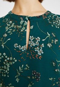 Blouse florale bleu sarcelle avec une ouverture en forme de goutte, fermeture par bouton au niveau du cou, et broderie détaillée de diverses plantes et fleurs.