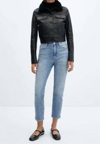 Chaqueta de cuero negra corta con cierre de botón y dos bolsillos en el pecho, llevada sobre un suéter negro de cuello alto, combinada con jeans de tiro alto de color azul claro y zapatos planos negros.