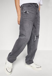 Šedé denimové kalhoty s popraskanými detaily a uvolněným střihem. Mají roztržení a kontrastní švy, doplněné bílými teniskami.