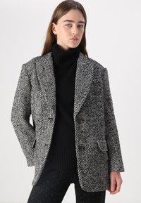 Blazer à motif pied-de-poule en noir et blanc avec revers, deux boutons et poches avant, porté par-dessus un col roulé noir.