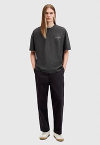 Tumši pelēka īsroku T-krekls ar apaļu apkakli, brīva piegriezuma un logotipa detaļa uz krūtīm, kopā ar melnām biksēm un baltām sporta kurpēm.