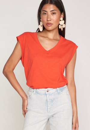 Donna con capelli lunghi e scuri che indossa una canotta arancione senza maniche con scollo a V, jeans azzurri a vita alta e orecchini beige vistosi.