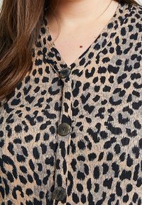 Leopardenmuster-Bluse in Beige und Schwarz, mit V-Ausschnitt, strukturiertem Stoff und zwei schwarzen Knöpfen entlang der Vorderseite.