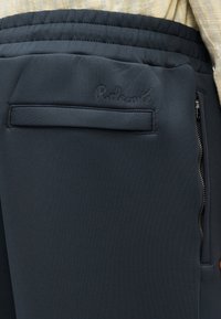 Pantalon de sport bleu foncé en tissu lisse et extensible. Comprend une poche zippée et un logo embossé sur la ceinture.