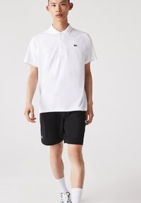 Vit polo skjorta i andningsbart material med en grön logotyp, parat med svarta shorts. Har korta ärmar och klassisk krage.