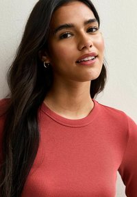 Jeune femme aux longs cheveux bruns portant de petites boucles d'oreilles en anneau et un haut à manches longues côtelé rouge, souriant doucement devant un mur uni.