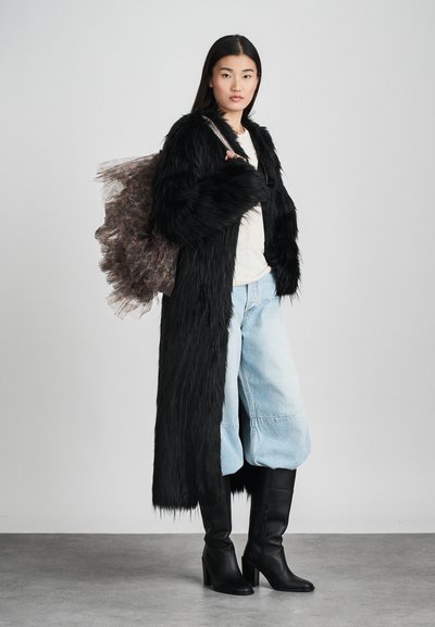 Schwarzer Kunstfellmantel, helles Hemd, weite hellblaue Jeanshose und knielange schwarze Absatzstiefel. Transparente, schichtweise Tasche mit dunklen Akzenten.