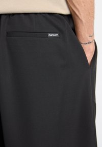 Black pants made from smooth fabric, featuring a back pocket with a stitched logo label. The design includes a relaxed fit and elastic waistband.