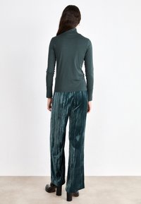 Dark green turtleneck top with long sleeves paired with pleated, shiny dark green wide-leg pants. Smooth texture with a fitted design.