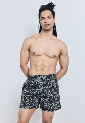 Homme torse nu avec des dreadlocks, debout sur un fond clair, portant un short de bain noir avec des motifs abstraits de lettres blanches.