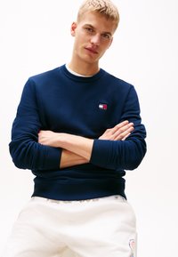 Marineblauwe sweatshirt van textuurstof met ribgebreide boorden en onderkant, voorzien van een klein rood en wit logo op de linkerborst. Gecombineerd met lichtgrijze broek.