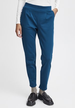 Türkisfarbene, tapered Hosen mit Falten an der Taille, aus weichem Stoff gefertigt. Kombiniert mit gestrickten Socken und schwarzen Slippern.