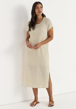 Kobieta o długich ciemnych włosach, ubrana w kremową dzianinową sukienkę midi z krótkimi rękawami i rozcięciem z boku, stojąca boso przy białej ścianie.