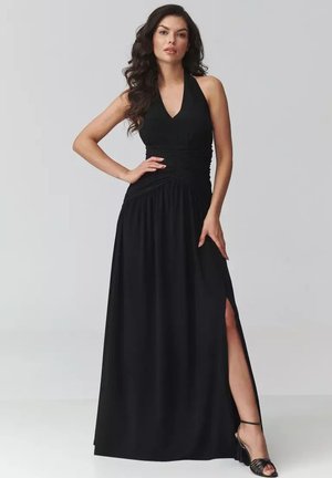 Kobieta ubrana w czarną, bezrękawnikową suknię na szyję halter z wysokim rozcięciem, pozująca z jedną ręką na biodrze na jednolitym szarym tle.