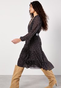 Schwarz gepunktetes Kleid aus durchsichtiger Stoff, mit langen Ärmeln und einem gestuften Rock. Getragen mit beigen knielangen Stiefeln und einem taillierten Gürtel.