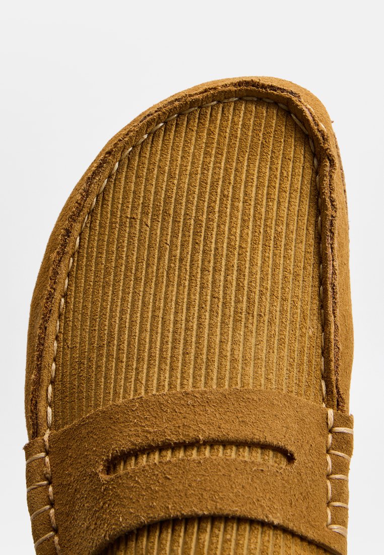 Mocassin en suede marron avec des bords cousus et une texture nervurée sur le dessus, présentant un détail de découpe horizontal sur la tige.