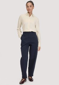 Vrouw die staat in een crèmekleurige knoopblouse, marineblauwe broek met hoge taille en zijzakken, en donkerbruine platte schoenen.