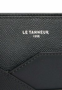 Portefeuille en cuir noir texturé avec des accents noirs lisses. Présente le logo en relief "LE TANNEUR 1898" en argent. Design compact.