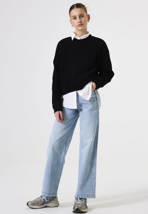 Jonge persoon in lichte spijkerbroek, een zwart sweatshirt over een wit shirt en zilveren hardloopschoenen tegen een effen achtergrond.