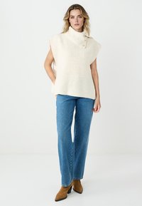 Top smanicato in maglia a coste color crema con collo alto e bottoni laterali, abbinato a jeans blu a gamba larga e scarpe tacchi marroni.